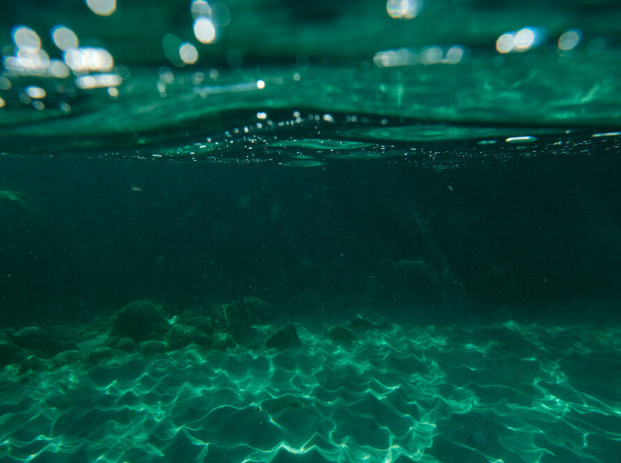 Water surface with light penetrating through the water, reaching the sandy sea bottom. On the sea bottom there are a few rocks.